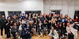Group photo of attendees at the Columbia Convening on AI Openness and Safety, smiling and waving while wearing blue, red, and white berets, seated and standing in a brightly lit room with large windows.